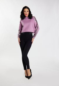 Pulover lila cu mâneci decorative din paiete, asortat cu colanți negri și tocuri negre ascuțite, pe un fundal neutru.
