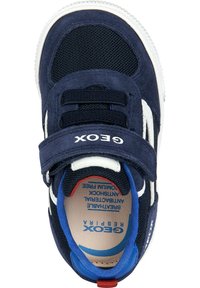 Zapatilla azul marino con parte superior de ante, paneles de malla y una correa blanca con marca. Presenta cierre de velcro y un interior acolchado.