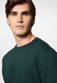 Ciemnozielona bluza z okrągłym dekoltem, charakteryzująca się miękką fakturą i przeszyciami, noszona przez modela o neutralnym wyrazie twarzy na jasnym tle.