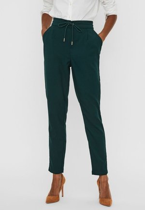 Pantalons verts à taper avec une taille élastique et un cordon de serrage, dotés de poches latérales et d'une texture lisse. Portés avec des talons hauts couleur tan.