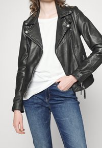 Kvinna som bär en svart läderjacka, vit skjorta och blå jeans med en hand i fickan mot en enfärgad bakgrund.