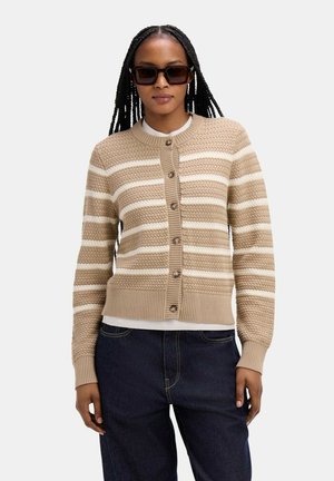 Femme portant un cardigan tricoté à rayures beige et blanc, un jean foncé et de grandes lunettes de soleil noires, sur un fond uni.