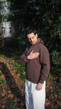 Fiatal férfi barna kapucnis pulóverben és fehér nadrágban áll a szabadban, kezét a mellkasára helyezi, csukott szemmel, zöld növények és lehullott levelek veszik körül.