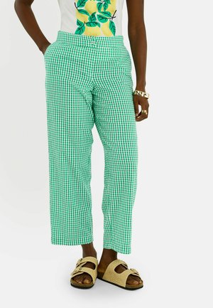 Personne portant un pantalon à carreaux verts et blancs, un haut blanc avec un imprimé citron et feuille, des sandales beiges et des bijoux dorés.