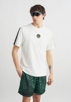 Giovane uomo che indossa una maglietta bianca Nike, pantaloncini verdi e neri con motivo, occhiali da sole neri e un tatuaggio sul braccio su uno sfondo uniforme.