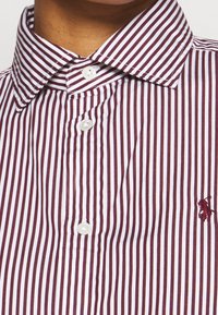Camisa de botones a rayas con rayas verticales en color burdeos y blanco, un cuello clásico y un pequeño logo bordado en el lado izquierdo del pecho.