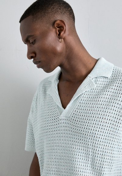 Polo de punto de manga corta en azul claro, con una textura de tejido abierto y un cuello plano. Presenta un diseño de escote en V profundo.