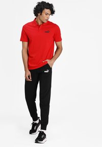 Maglietta polo rossa con logo Puma nero, abbinata a pantaloni jogger neri con logo Puma bianco. Scarpe da ginnastica nere completano il look.