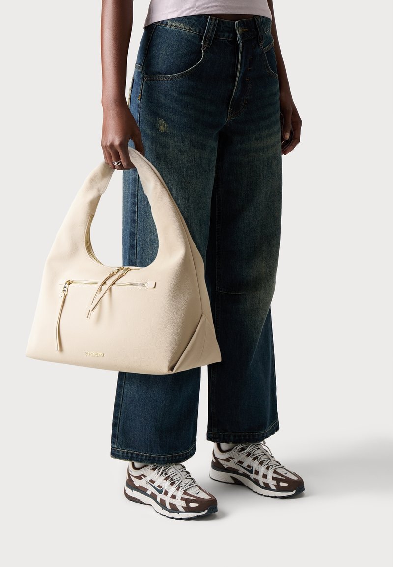Personne tenant un grand sac à main en cuir beige, portant un jean large foncé et des baskets marron et blanches sur un fond clair.