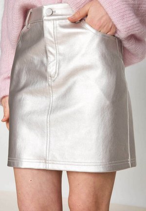 Vrouw in een glanzende zilveren leren minirok met haar hand in de voorzak en een roze zachte trui.