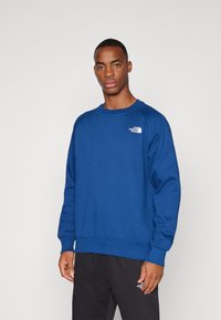 Sudadera de forro polar azul con cuello redondo y puños acanalados, con un pequeño logo blanco en la parte superior izquierda del pecho y sin estampados.