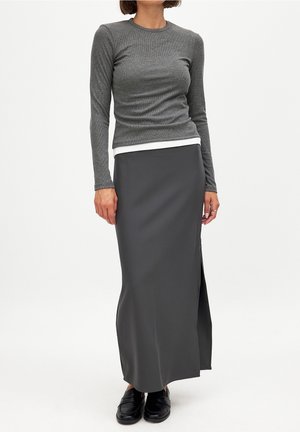 Femme portant un haut moulant à manches longues couleur charbon superposé à un haut blanc, associé à une longue jupe gris foncé avec une fente latérale et des mocassins noirs.