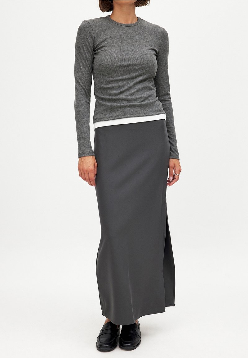 Femme portant un haut moulant à manches longues couleur charbon superposé à un haut blanc, associé à une longue jupe gris foncé avec une fente latérale et des mocassins noirs.