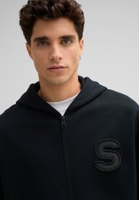 Haut à zip noir en tissu doux, avec un logo 'S' texturé sur la poitrine et une capuche avec cordon de serrage. Le modèle fait face à l'avant.