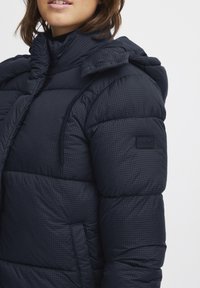Marineblauwe pufferjack met een geruit patroon, voorzien van een hoge kraag, een capuchon en een textuurstof met een label op de mouw.