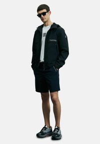 Veste noire imperméable avec capuche, zip frontal et poches latérales. Portée par-dessus une chemise blanche et associée à un short sombre et des baskets grises.
