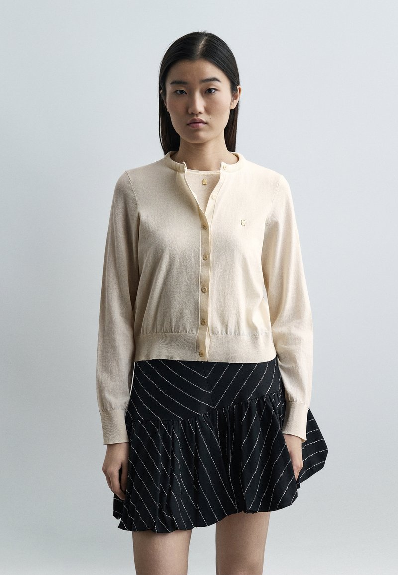 Γυναίκα με ίσια σκουρόχρωμα μαλλιά που φοράει μπεζ ζακέτα με κουμπιά και μαύρη φούστα με λευκές διαγώνιες ρίγες, όρθια μπροστά σε απλό φωτεινό φόντο.