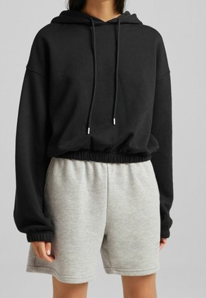 Osoba v černé crop mikině s kapucí na stahovací šňůrku a světle šedých šortkách s elastickým pasem na neutrálním pozadí.