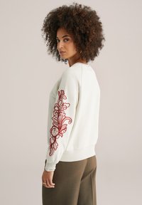 Felpa color crema con intricati ricami floreali rossi sulla manica sinistra, polsini aderenti e scollo rotondo.