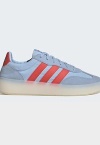 Zapatilla azul clara con tres rayas rojas, detalles de ante azul claro, una suela gruesa translúcida y cordones sobre un fondo liso.
