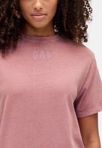 Brudnoróżowy t-shirt z okrągłym dekoltem, z subtelnym jasnoszarym logo "GAP" na piersi, wykonany z miękkiego, strukturalnego materiału, z krótkimi rękawami.