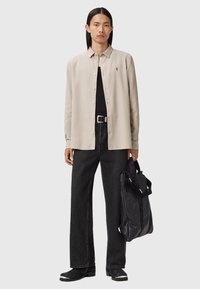 Camicia beige a bottoni, t-shirt nera, pantaloni jeans neri a gamba larga, stivali neri con dettagli bianchi e una borsa a tracolla nera con logo.