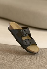 Sandal neagră cu două catarame, având un suport din plută, talpă din cauciuc texturat și partea superioară din material sintetic neted. Accente cu catarame metalice.