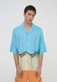 Camicetta azzurra chiaro a maniche corte con un design a bottoni. La parte inferiore presenta una stampa colorata di case stilizzate.