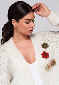 Vit stickad kofta med ribbstickade kanter, prydd med dekorativa blomapplikationer i röd, grön och beige. Modellen bär stora hoops.