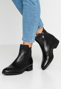 Bottines en cuir noir avec une finition lisse, des panneaux latéraux élastiques et un petit talon carré. Elles disposent d'une fermeture à glissière avec un accent circulaire.