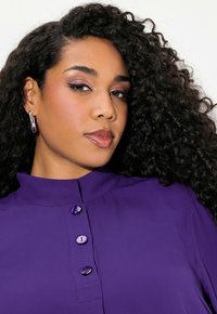 Fialová blůza s vysokým límcem, se třemi fialovými knoflíky. Tkanina je hladká a design zahrnuje upravený střih.