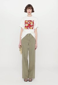 Weißes übergroßes T-Shirt mit rotem Blumenmotiv, kombiniert mit lockeren olivgrünen Hosen und schwarzen Riemchensandalen. Die Modells hält eine goldene Clutch.