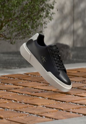 Sneaker Calvin Klein bianco e nero con suola bianca sospesa sopra un pavimento esterno in legno e ciottoli, con sfondo sfocato di muro e vegetazione.