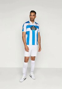 Muž v modro-bielom pruhovanom futbalovom drese s bielymi šortkami, vysokými bielymi ponožkami a bielymi kopačkami stojí proti jednému pozadiu.