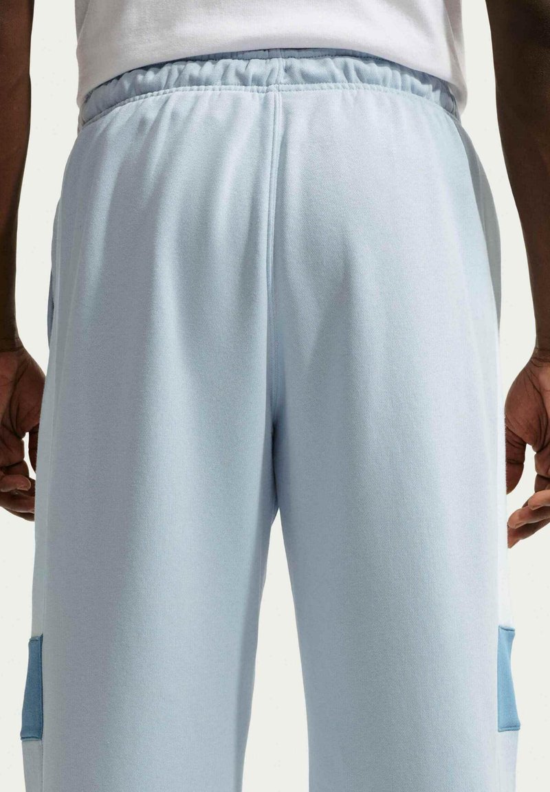 Persoon staat met de armen langs het lichaam, draagt lichtblauwe joggingbroek en een wit shirt, vanaf de achterkant op taillehoogte getoond.