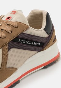 Sneaker mit einem oberen Teil aus sandfarbenem Wildleder und beigem Netzgewebe, schwarzen Akzenten, lila Branding und einer orangefarbene Sohle mit strukturierten Details.