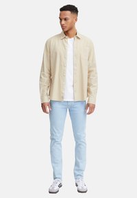 Helles beiges Knopfhemd über einem weißen T-Shirt, kombiniert mit hellblauen Jeans und grau-weißen Sneakers. Einfaches Design mit einer Brusttasche.