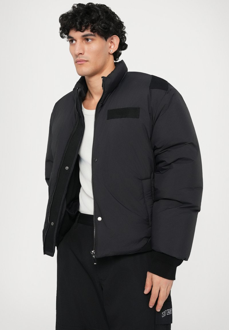 Schwarze Daunenjacke mit hohem Kragen, vorderem Druckverschluss und Klettverschlussakzenten, mit glatter Textur und strukturiertem Schnitt.