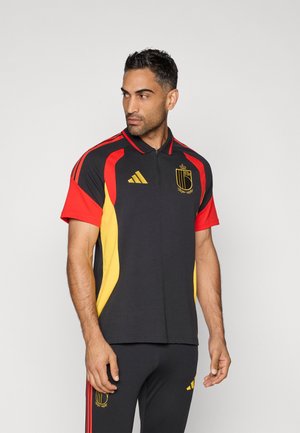 Homem vestido com polo de futebol da Bélgica nas cores preto, vermelho e amarelo, acompanhado de calças a condizer, de pé contra um fundo cinza claro e simples.