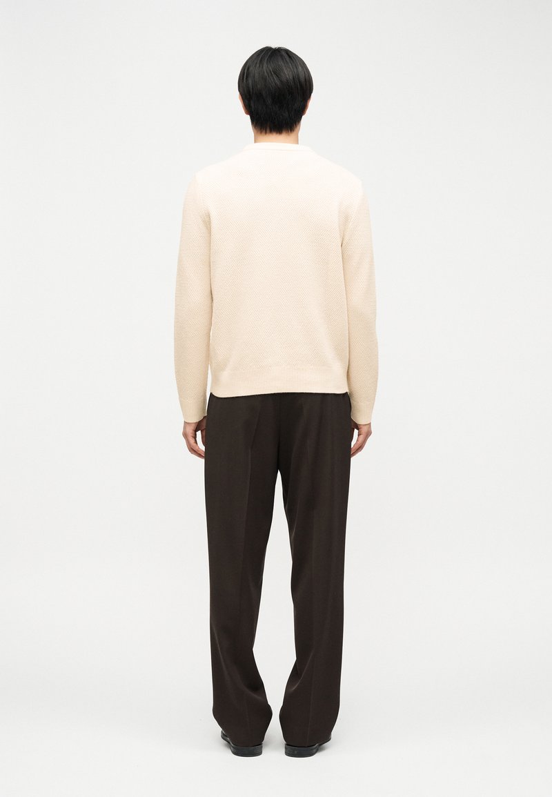 Person standing, facing away, wearing a cream textured sweater, dark loose trousers, and black shoes on a plain white background.