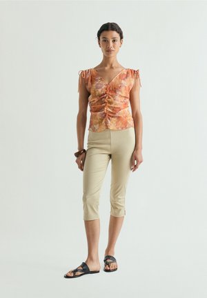 Vrouw staand in beige cropped broek, oranje bloemen gerimpelde V-hals top, zwarte sandalen en een bruine armband aan de linkerpols tegen een effen achtergrond.