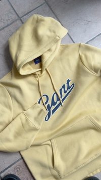 Sweatshirt amarelo com capuz e um grande logótipo azul marinho "Gant". Tem um bolso canguru e punhos canelados. Textura de tecido macio.