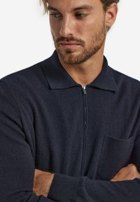 Marinblå zip-jacka med krage, ribbade muddar, bröstficka och ett mjukt, texturerat tyg. Har en subtil glans och minimalistisk design.