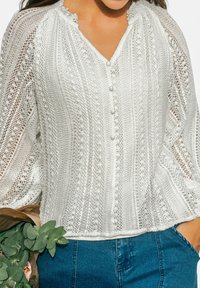 Blusa bianca a maglia con maniche lunghe e bottoni, indossata con jeans blu, mentre tiene delle foglie di eucalipto verdi avvolte in carta marrone.