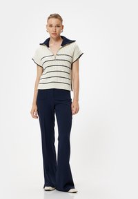 Stickad polo topp i krämfärg med marinblå ränder, med en dragkedja vid kragen. Matchas med mörk marinblå utsvängda byxor, i slätt material.