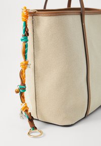 Beige tote bag with a textured surface, brown leather accents, and a colorful, braided strap featuring assorted knots and a keyring.
