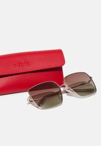Lunettes de soleil avec une monture argentée métallique et des verres marron dégradés, accompagnées d'une étui en cuir rouge texturé embossé avec "GUESS".