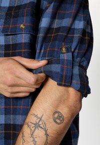 Blue and black plaid flannel shirt with orange accents, featuring a buttoned sleeve and visible stitching. Hand adjust the cuff, showing texture.