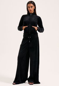 Zwart velours trainingspak met een zipjack, voorzien van zakken, en lange, wijde broek met een elastische tailleband. Overal een gladde textuur.