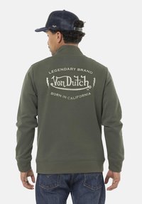 Homme portant une veste olive de la marque Von Dutch avec un logo et le texte "Legendary Brand Born in California" au dos, un jean sombre et une casquette marine.